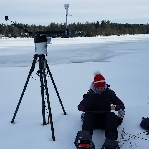 Dr. Jenna Robinson downloading data from a weatherstation on the shore of a frozen Lake Waynewood.